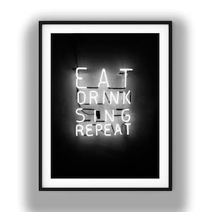 May include: A framed wall art print featuring a black background with white neon-style lettering that reads EAT DRINK SING REPEAT. The text is arranged in four stacked lines, creating a glowing effect against the dark, solid-colored backdrop.