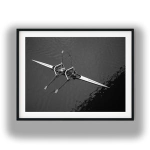 May include: A black and white aerial photograph of a long, narrow rowing shell on dark water. Two rowers are positioned in the center of the boat, each operating a pair of oars that extend outward, creating a symmetrical shape against the water.