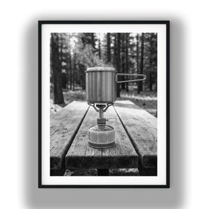 May include: Black and white photograph of a camping stove with a pot on a wooden picnic table. Steam rises from the pot, suggesting it is in use. The background features a forest with tall trees.