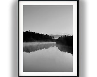 May include: A black and white framed wall art print featuring a serene landscape of a calm river reflecting a dense forest line under a soft, misty sky. The composition captures the quiet atmosphere of early morning with dark silhouettes.