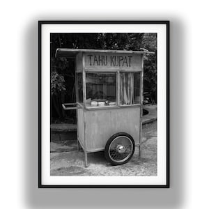 May include: A black and white photograph of a street food cart with a metal frame, a single spoked wheel, and a glass-enclosed display area. The words TAHU KUPAT are printed in dark letters across the top front panel of the rectangular cart structure.