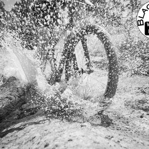 May include: A black and white action photograph of a bicycle wheel splashing through a muddy puddle, creating a large spray of water droplets. In the upper right corner, a circular logo features a camera icon with the text BLACK AND WHITE LENS.