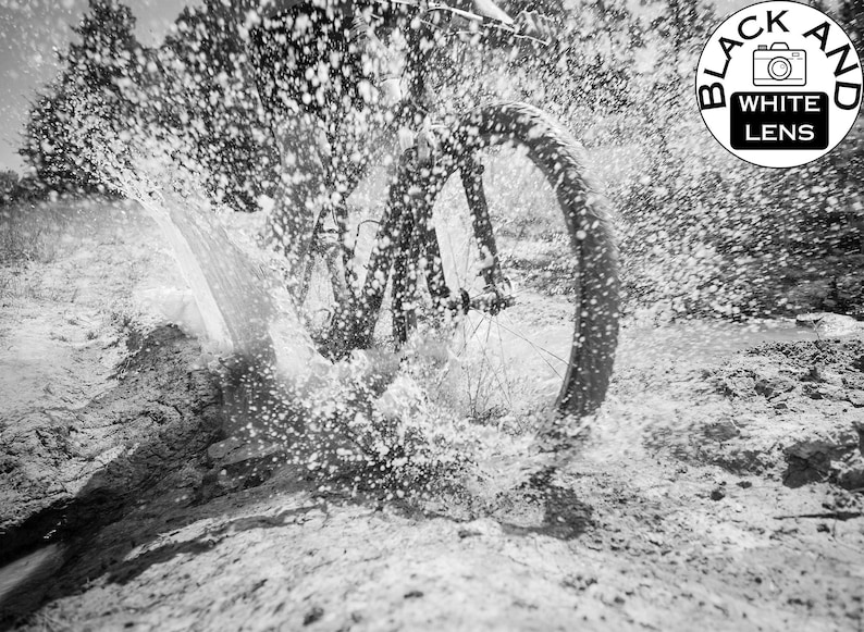 May include: A black and white action photograph of a bicycle wheel splashing through a muddy puddle, creating a large spray of water droplets. In the upper right corner, a circular logo features a camera icon with the text BLACK AND WHITE LENS.