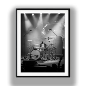 May include: A black and white photograph of a drum kit set up on a stage. The kit includes a bass drum, snare, toms, and cymbals mounted on stands. Two microphone stands are positioned on either side of the drums under bright stage lights.