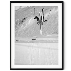 May include: A black and white photograph of a skier performing a backflip in mid-air. The skier is wearing a ski suit and has their skis crossed. The skier is against a snowy background.