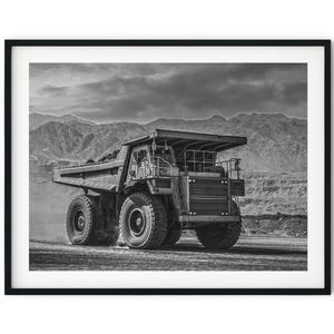 Könnte beinhalten: Ein großes Schwarzweißfoto eines großen, grauen Muldenkippers, der auf einer unbefestigten Straße in einer Wüstenlandschaft fährt. Der LKW transportiert eine Ladung Erde oder Gestein. Der Hintergrund ist eine bergige Wüstenlandschaft.