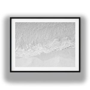 Puede incluir: Una impresión de arte mural enmarcada en blanco y negro que presenta una vista aérea de las olas del océano bañando suavemente una playa de arena. La composición captura el contraste entre el agua texturizada y espumosa y la orilla clara.