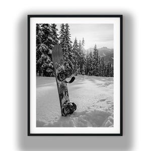 Puede incluir: Fotografía enmarcada en blanco y negro de una tabla de snowboard apoyada en la nieve profunda. La tabla, con fijaciones negras, se encuentra frente a un paisaje montañoso nevado con árboles de hoja perenne. El sol brilla en el fondo.