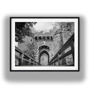 May include: A black and white photograph of a stone castle gatehouse with a wooden bridge leading to the arched entrance. The structure features weathered masonry, crenelated battlements, and a cloudy sky, framed by a dark border.