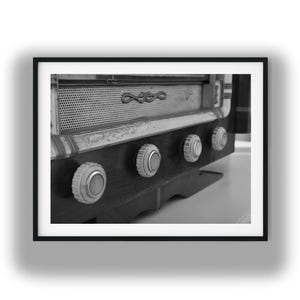 Può includere: Una fotografia in bianco e nero di una radio vintage caratterizzata da una griglia per altoparlanti in metallo perforato con un emblema decorativo, una base in legno scuro e quattro manopole circolari in una cornice nera.