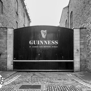 May include: A black and white photograph of a large metal gate with the words "GUINNESS ST. JAMES'S GATE BREWERY, DUBLIN" on it. The gate is situated in a brick alleyway.