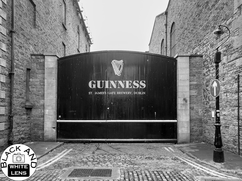 May include: A black and white photograph of a large metal gate with the words "GUINNESS ST. JAMES'S GATE BREWERY, DUBLIN" on it. The gate is situated in a brick alleyway.