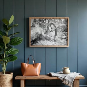 May include: A framed black and white photograph depicting a bicycle wheel splashing through water, displayed above a wooden bench holding a tan leather tote bag, a small ceramic pitcher, and a folded grey textured throw blanket.