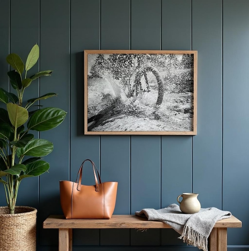 May include: A framed black and white photograph depicting a bicycle wheel splashing through water, displayed above a wooden bench holding a tan leather tote bag, a small ceramic pitcher, and a folded grey textured throw blanket.