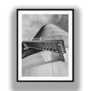 May include: Black and white photograph of a tall, cylindrical structure with a sloped roof and a metal staircase. The image is framed in a black border, with a cloudy sky in the background. The structure appears to be made of concrete.