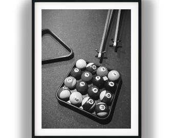 May include: A black and white framed print featuring a square rack filled with numbered billiard balls, two pool cues with bridge heads resting nearby, and a triangular rack frame positioned on a textured surface, capturing a game setup.