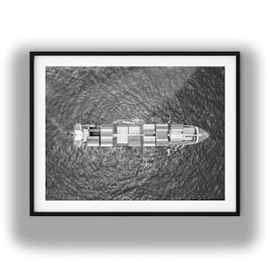May include: A black and white aerial photograph of a large cargo ship navigating through open water. The vessel is loaded with numerous rectangular shipping containers arranged in rows on its deck, creating a stark contrast against the textured, rippling surface of the dark ocean water surrounding the ship.