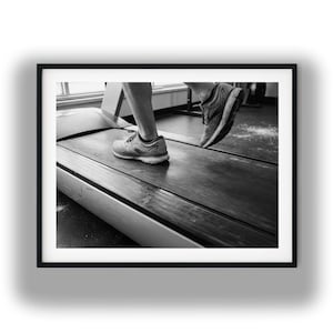 May include: Black and white image of a person running on a treadmill. The person's legs and feet are visible, wearing running shoes. The treadmill is in motion, with the belt moving. The background is blurred, suggesting an indoor gym setting.