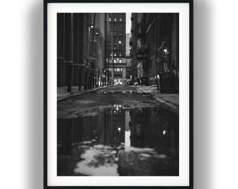 May include: A black and white framed photograph of a narrow city alleyway at night. The scene features tall buildings on both sides, a wet asphalt ground with a large puddle reflecting the urban architecture, and distant street lights glowing.