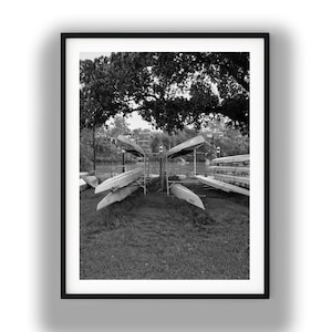 Może przedstawiać: Czarno-biała fotografia kilku kajaków i kajaków przechowywanych na stojakach w pobliżu zbiornika wodnego. Łodzie ułożone są wachlarzowato, z dużym drzewem w tle. Obraz jest oprawiony w czarną ramkę.