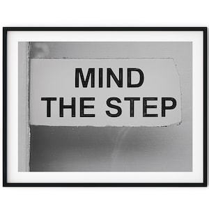 May include: Framed black and white print displaying the words "MIND THE STEP" in a bold, sans-serif font. The sign is rectangular with a slightly torn edge, set against a light grey background. The print is enclosed within a black frame.