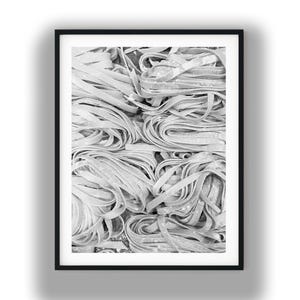 May include: Black and white photograph of fresh, uncooked pasta. The image features a close-up view of tagliatelle noodles, arranged in a dense, textured pattern. The pasta is pale in color, with a slightly rough surface.