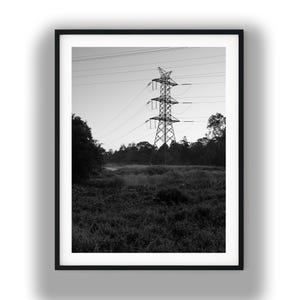May include: A black and white framed photograph featuring a tall metal electrical transmission tower standing in a field. The structure is centered against a clear sky, with power lines extending horizontally across the frame above trees.