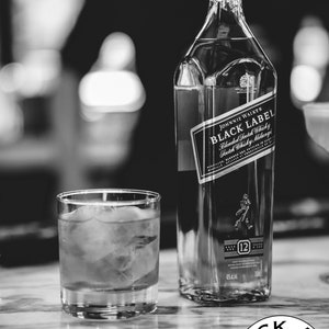 May include: A black and white photograph of a square glass bottle of Johnnie Walker Black Label Blended Scotch Whisky next to a short glass filled with liquid and ice cubes. The bottle label displays the brand name and 12-year age statement.