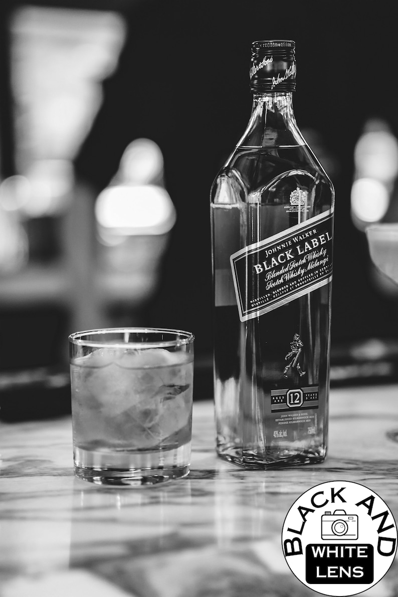 May include: A black and white photograph of a square glass bottle of Johnnie Walker Black Label Blended Scotch Whisky next to a short glass filled with liquid and ice cubes. The bottle label displays the brand name and 12-year age statement.
