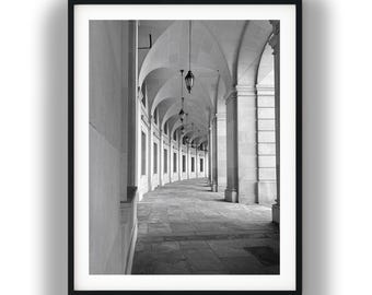 May include: A black and white framed photograph showing a long, curved stone arcade with a series of arched columns and hanging pendant lights. The perspective leads the eye down a paved walkway under a vaulted ceiling, creating a sense of depth.