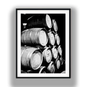May include: Black and white photograph of a stack of wooden wine barrels. The barrels are arranged in a pyramid shape, with several rows of barrels visible. The image is framed in a black border, and the background is dark.