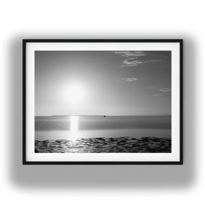 Può includere: Fotografia in bianco e nero incorniciata di un paesaggio marino. Il sole splende luminoso, riflettendosi sull'acqua. Il primo piano mostra una costa strutturata. L'immagine è un paesaggio monocromatico.