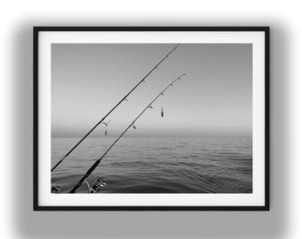 May include: A black and white framed photograph featuring two fishing rods extending diagonally over a calm, expansive body of water. The rods are equipped with reels and lines, set against a horizon line where the sky meets the sea.