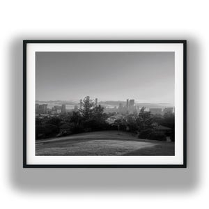 May include: Black and white framed photograph of a cityscape. The image features a grassy hill in the foreground, with trees and buildings in the mid-ground, and a skyline of skyscrapers in the distance. The sky is overcast.