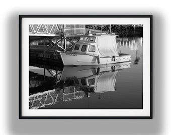 May include: Black and white framed photograph of a small boat named "Little Toot" docked at a pier. The boat is reflected in the calm water, with a white canopy and cabin. The pier structure is visible in the background.