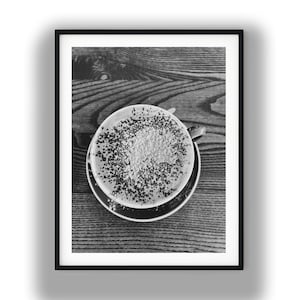 May include: A black and white photograph of a coffee cup filled with a beverage topped with foam and a dusting of dark powder. The cup sits on a matching saucer, and the entire arrangement is framed within a simple black border.
