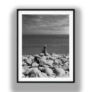 May include: Monochrome framed print depicting a coastal landscape. A cairn of stones stands on a pebble beach, with the sea and a cloudy sky in the background. The print is presented in a black frame.
