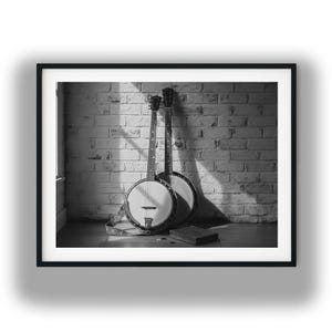 May include: Black and white photograph of two banjos leaning against a brick wall. The banjos have round bodies and long necks. A small wooden box and a few picks are on the floor in front of them. The image is framed in black.