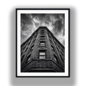 May include: Black and white photograph of a tall brick building with multiple windows, framed in a black border. The perspective is from the ground looking up, with a cloudy sky above. The building's architecture is the central focus.