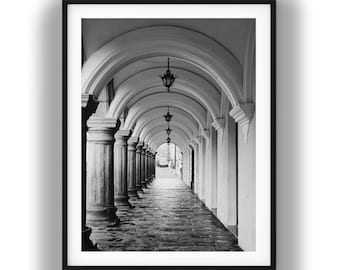 May include: A black and white framed photograph depicting a long stone colonnade with repeating arches. Hanging lanterns are suspended from the center of each archway, leading the eye toward a bright opening at the end of the corridor.