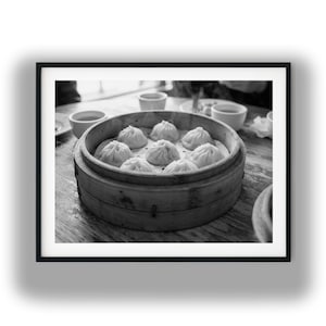Può includere: Una fotografia in bianco e nero di un cestello rotondo in bambù per la cottura a vapore contenente otto ravioli al vapore. I ravioli sono disposti in modo circolare all'interno del contenitore in legno, posato su un tavolo con ciotole bianche.