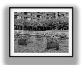 May include: Black and white photograph of an outdoor cafe scene. Small round tables and chairs are arranged on a brick patio. Wooden planters with greenery are in the foreground. Buildings and trees are in the background.