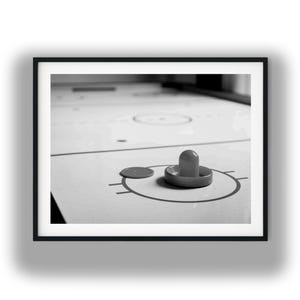 May include: A black and white photograph of an air hockey table featuring a smooth white surface with black circular markings and a center line. A single round puck and a handheld mallet are positioned on the table within the marked goal area.