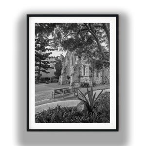 May include: A black and white framed photograph of a stone church building with arched windows, surrounded by mature trees. In the foreground, two wooden benches sit on a grassy lawn near a paved path, with a large spiky plant in the lower right.