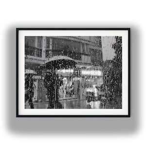 Könnte beinhalten: Schwarz-weiß gerahmte Fotografie einer verregneten Straßenszene. Regentropfen ziehen über das Glas und verdecken die Sicht auf Gebäude und Menschen. Ein beleuchtetes Schild ist in der Ferne zu sehen.