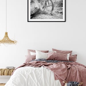 May include: A bedroom scene featuring a bed with white and dusty rose bedding, a woven pendant light, and a black-framed wall art print depicting a black and white photograph of a bicycle wheel splashing through water in a natural outdoor setting.