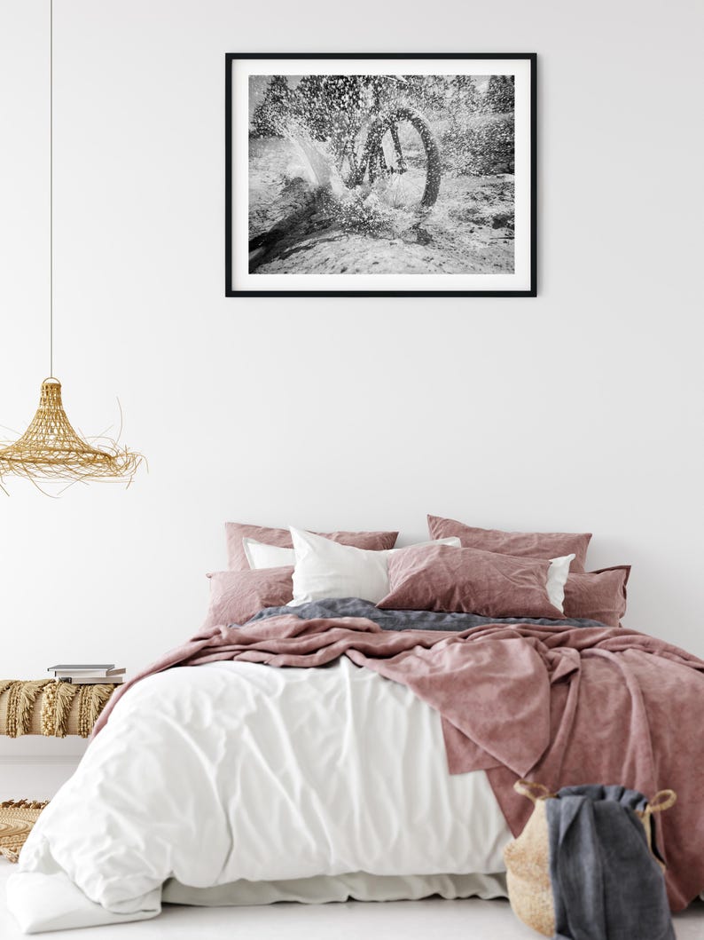 May include: A bedroom scene featuring a bed with white and dusty rose bedding, a woven pendant light, and a black-framed wall art print depicting a black and white photograph of a bicycle wheel splashing through water in a natural outdoor setting.