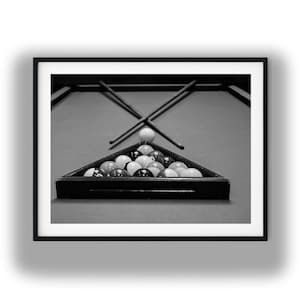 May include: A black and white framed print featuring a triangular rack filled with billiard balls on a pool table. Two crossed pool cues are positioned behind the rack, with a single white cue ball resting between the cues and the rack.