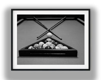 May include: A black and white framed print featuring a triangular rack filled with billiard balls on a pool table. Two crossed pool cues are positioned behind the rack, with a single white cue ball resting between the cues and the rack.