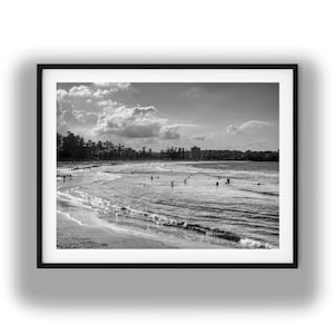 Könnte beinhalten: Schwarz-weiß gerahmte Fotografie einer Strandszene. Das Bild zeigt einen breiten Strand mit Menschen im Wasser und am Sand. Der Himmel ist mit Wolken gefüllt, und die Meereswellen sind sichtbar. Die Fotografie befindet sich in einem schwarzen Rahmen.